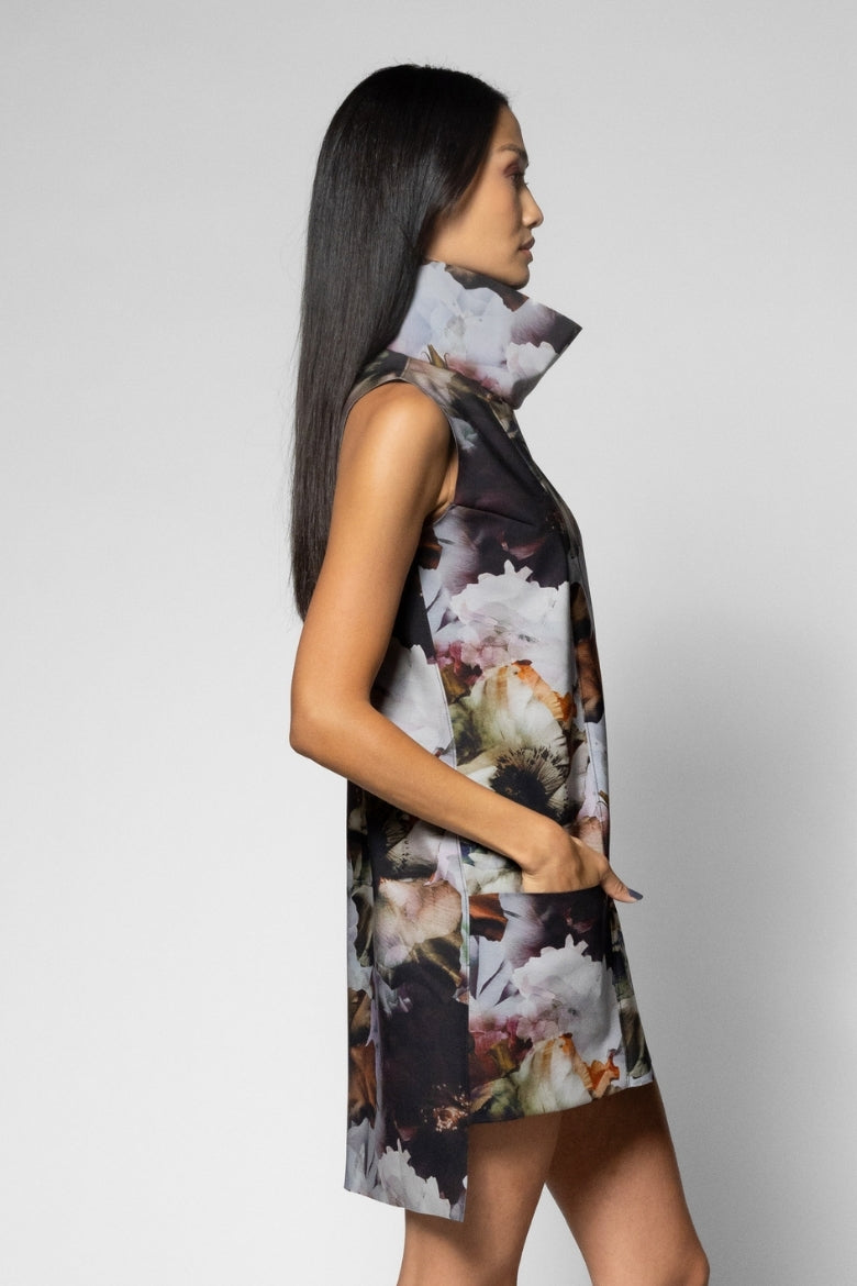 A woman shown in side profile wearing a sleeveless high-neck dress with a large-scale floral print in black, white, and soft peach tones, photographed against a light gray studio background.