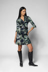 A woman modeling a short wrap-style dress with a dark green, black, and gray floral or watercolor-like print, cinched at the waist with a black belt, paired with black knee-high boots against a light gray studio background.