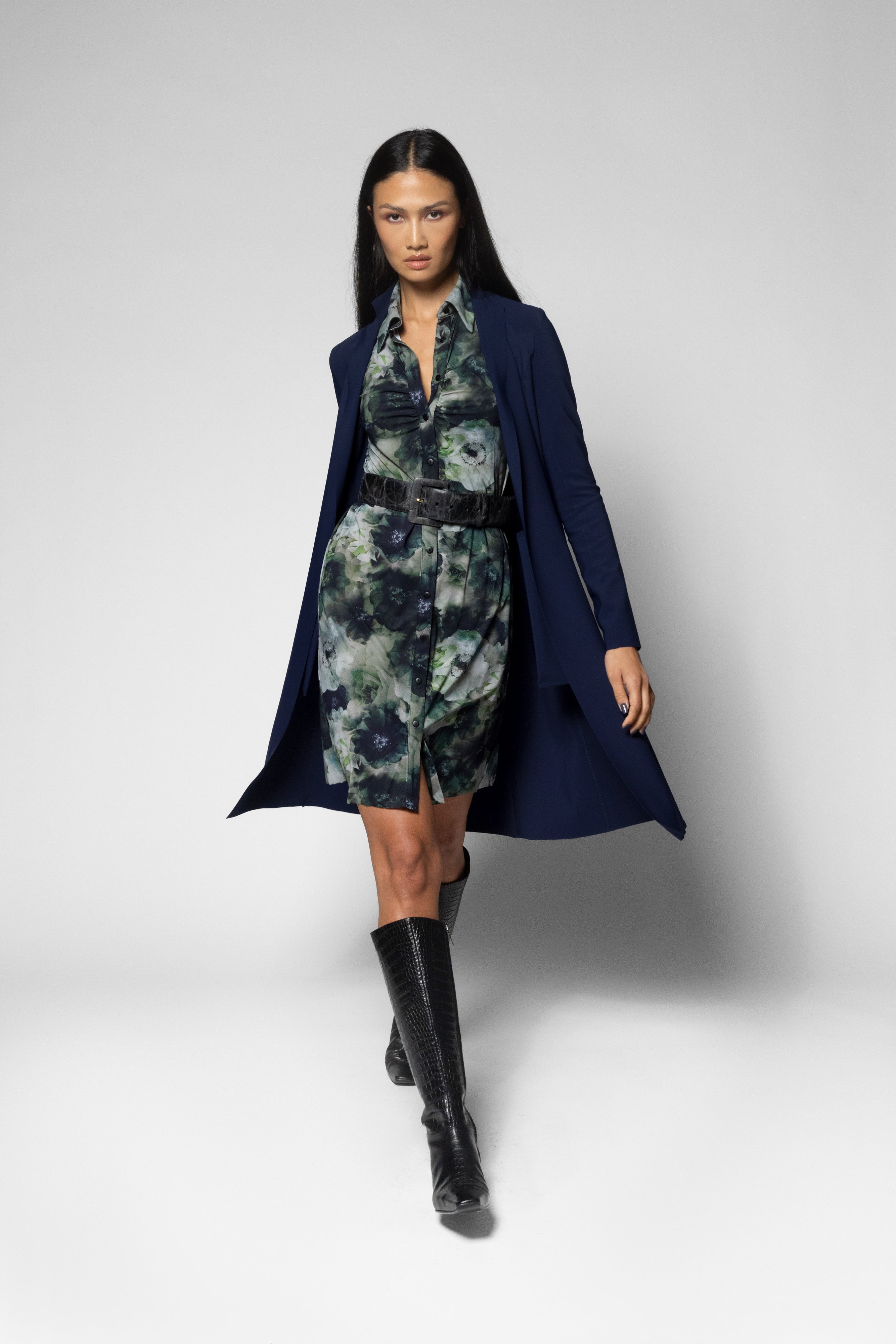A model walks against a light gray studio background wearing a navy blue open-front coat over a green and gray floral print dress with a black belt, paired with black knee-high boots.