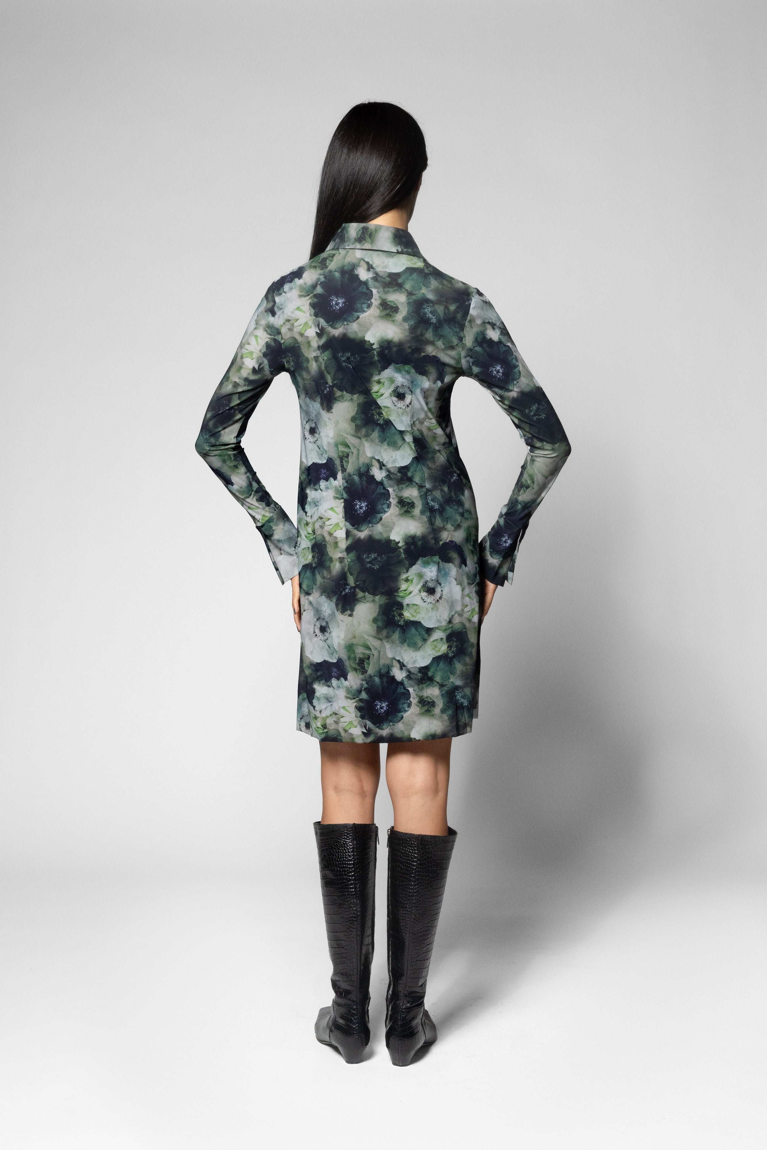 A model viewed from the back wearing a long-sleeve collared dress with a dark floral watercolor pattern in navy, green, and gray, paired with black knee-high boots, against a light gray studio background.
