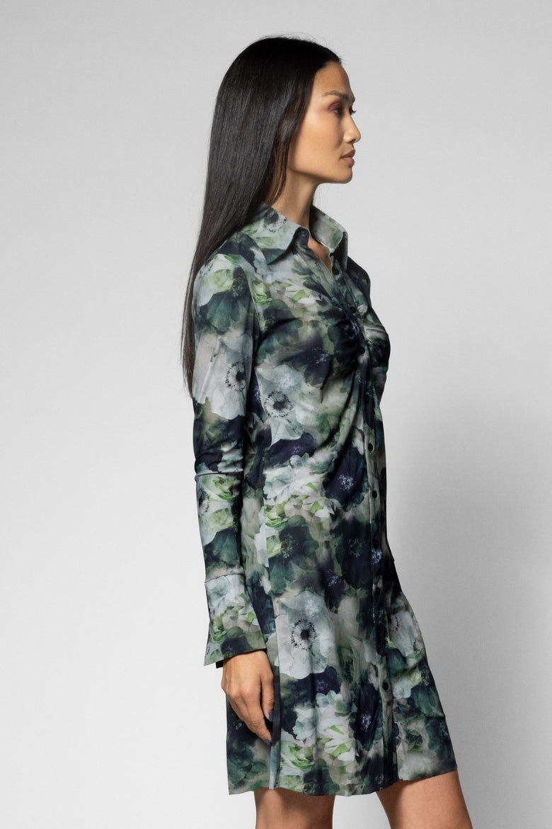 A woman modeling a long-sleeve button-front shirt dress with a green, gray, and navy floral watercolor print, shown from a side profile against a light gray studio background.