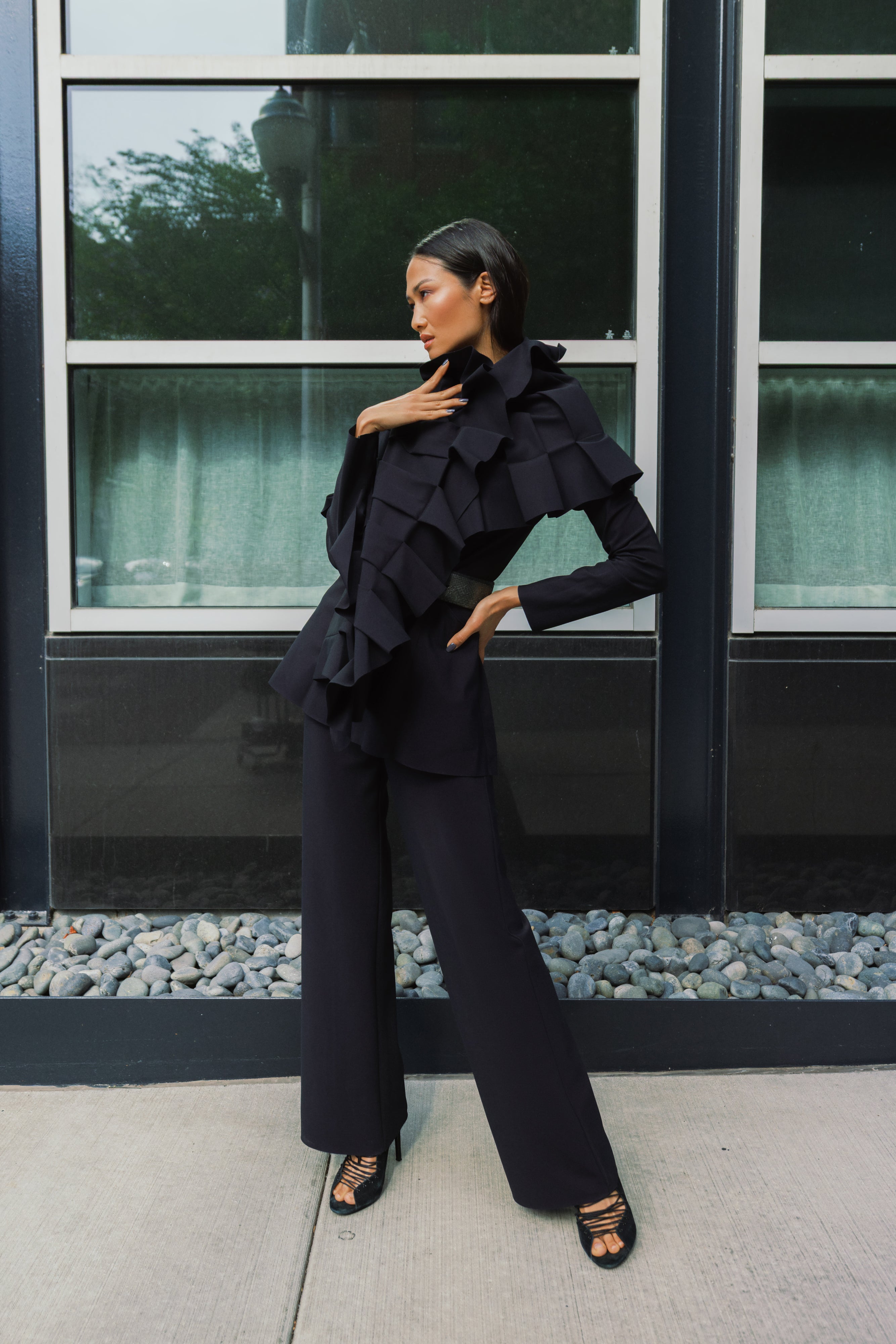 A woman models a black ruffled scarf over black jacket and matching wide-leg trousers, styled with black strappy heels, standing in front of a modern building facade with large windows.
