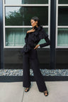 A woman models a black ruffled scarf over black jacket and matching wide-leg trousers, styled with black strappy heels, standing in front of a modern building facade with large windows.