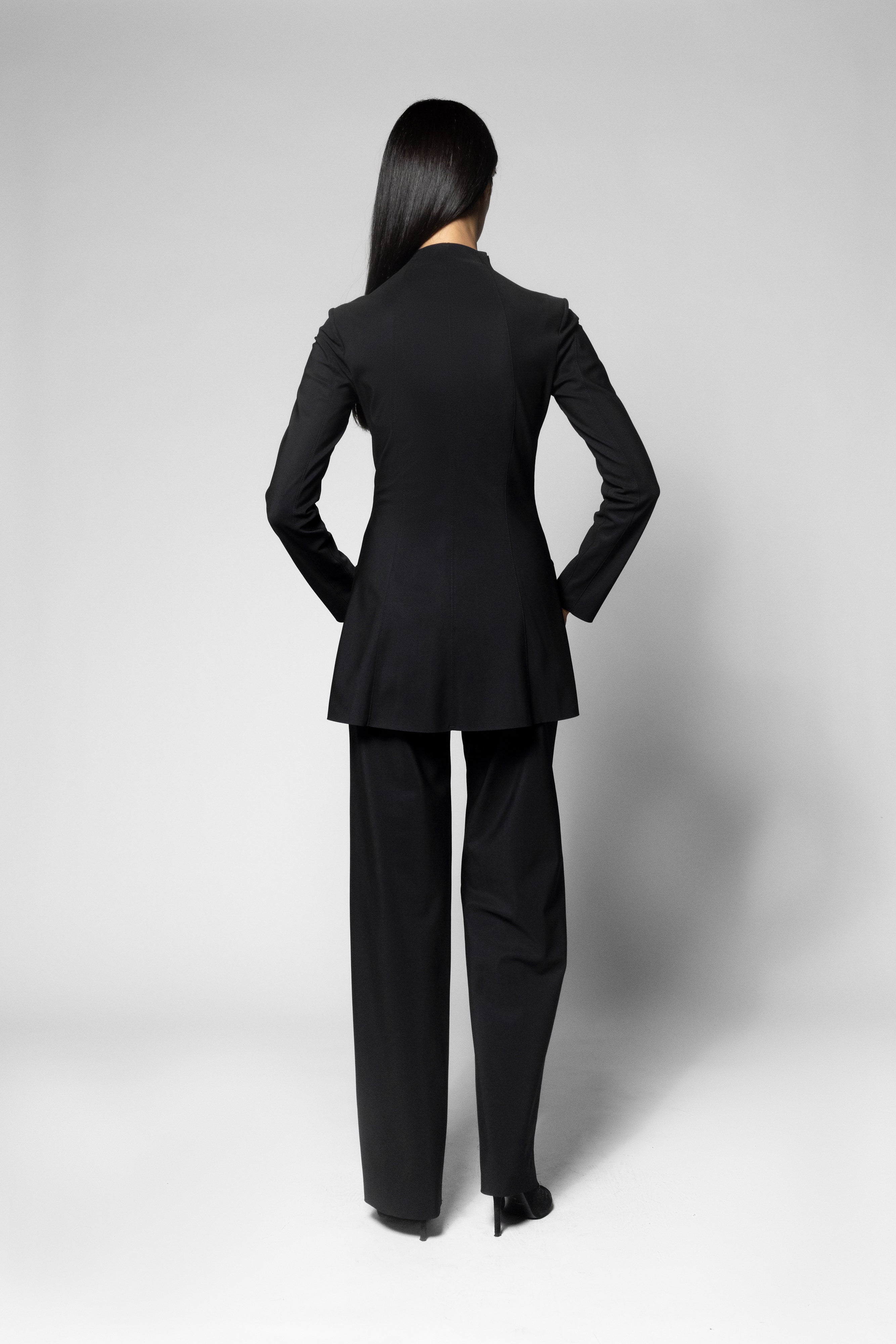 A woman viewed from the back wearing a tailored black jacket and matching wide-leg trousers against a light gray studio background.