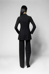 A woman viewed from the back wearing a tailored black jacket and matching wide-leg trousers against a light gray studio background.