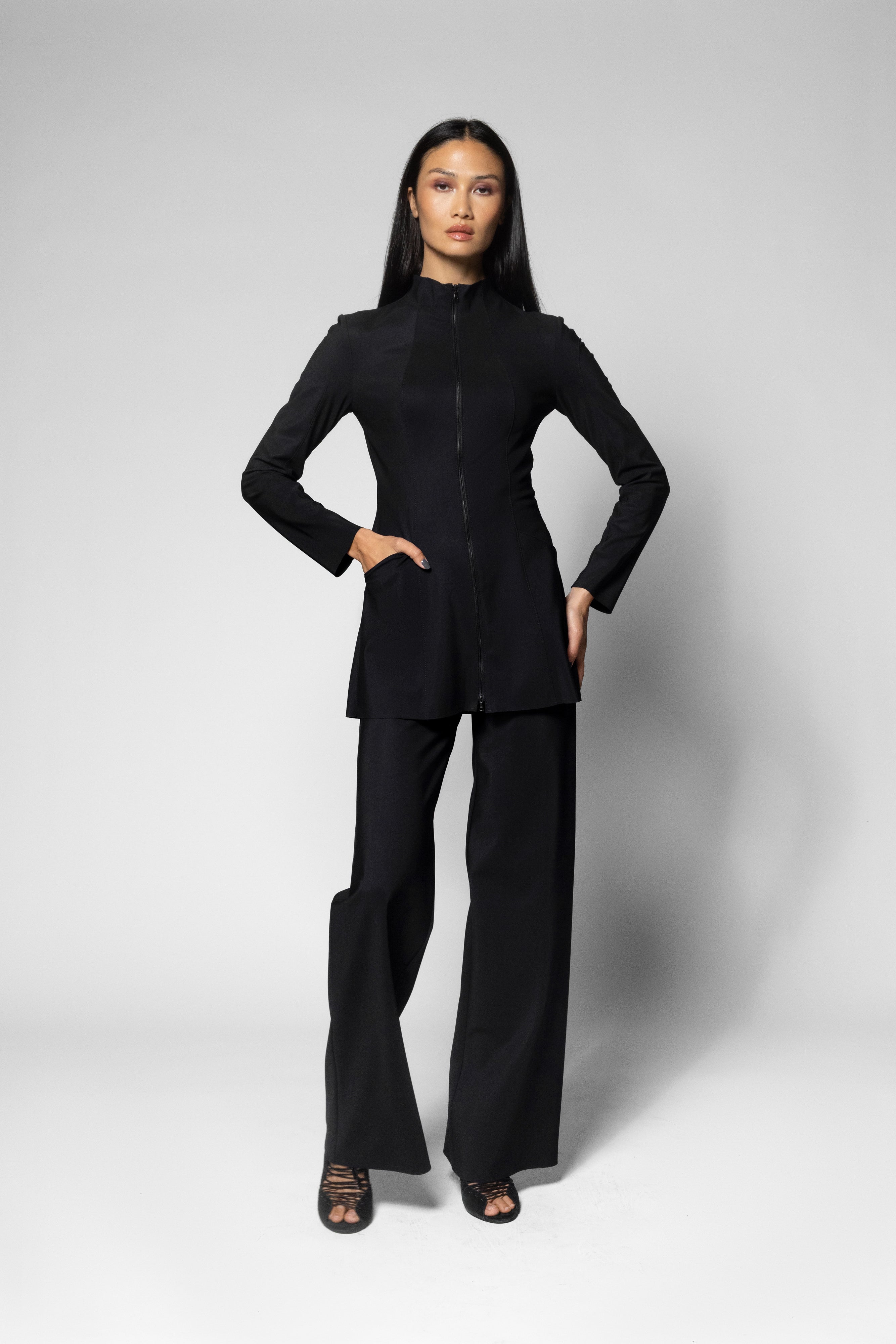 A model stands against a light gray studio background wearing a black long-sleeve jacket with a high mock neck and front zipper, paired with matching black wide-leg pants.
