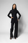 A model stands against a light gray studio background wearing a black long-sleeve jacket with a high mock neck and front zipper, paired with matching black wide-leg pants.