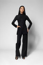 A model stands against a light gray studio background wearing a black long-sleeve jacket with a high mock neck and front zipper, paired with matching black wide-leg pants.