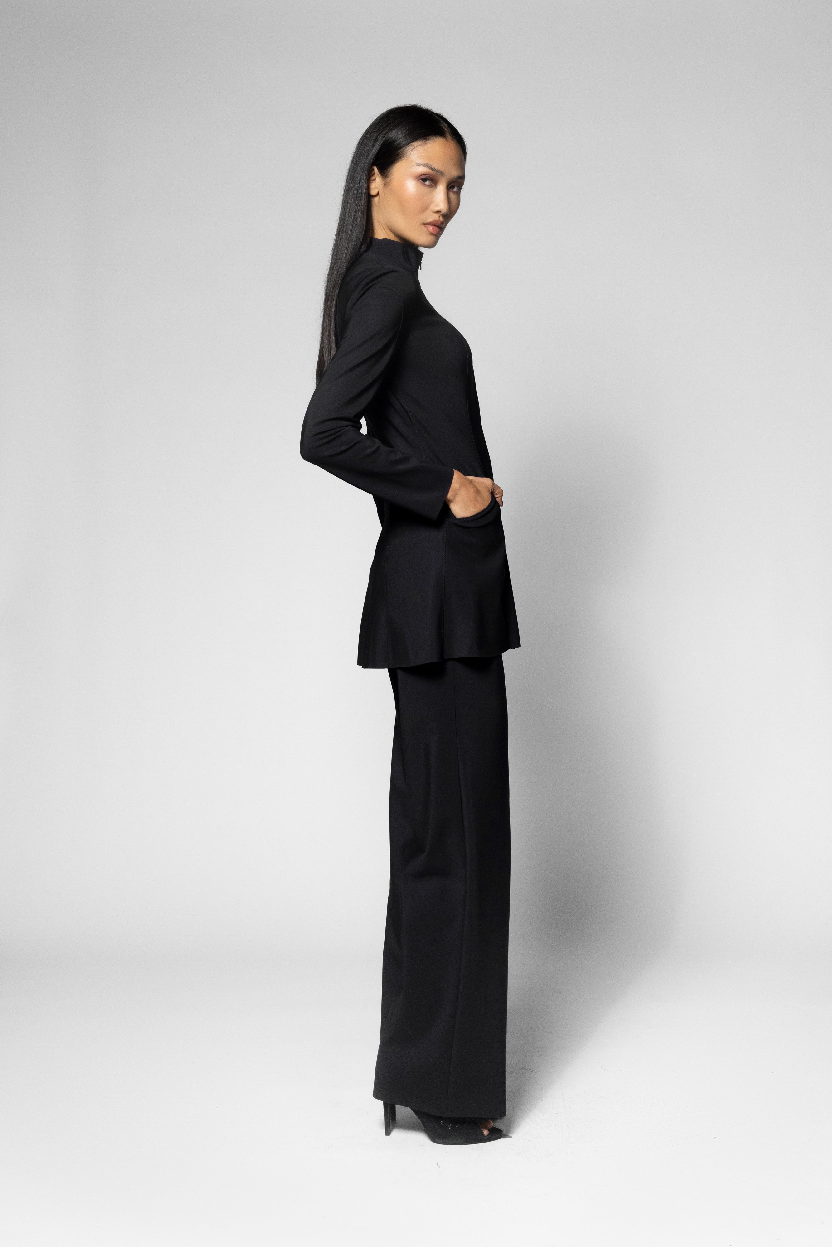 A woman modeling a black long-sleeve blazer with a peplum waist and matching wide-leg trousers, shown in profile against a light gray studio background.