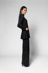 A woman modeling a black long-sleeve blazer with a peplum waist and matching wide-leg trousers, shown in profile against a light gray studio background.