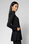 A woman with long dark hair is shown in profile wearing a black long-sleeve mock-neck jacket with a peplum hem and side pockets, paired with black pants, against a light gray studio background.