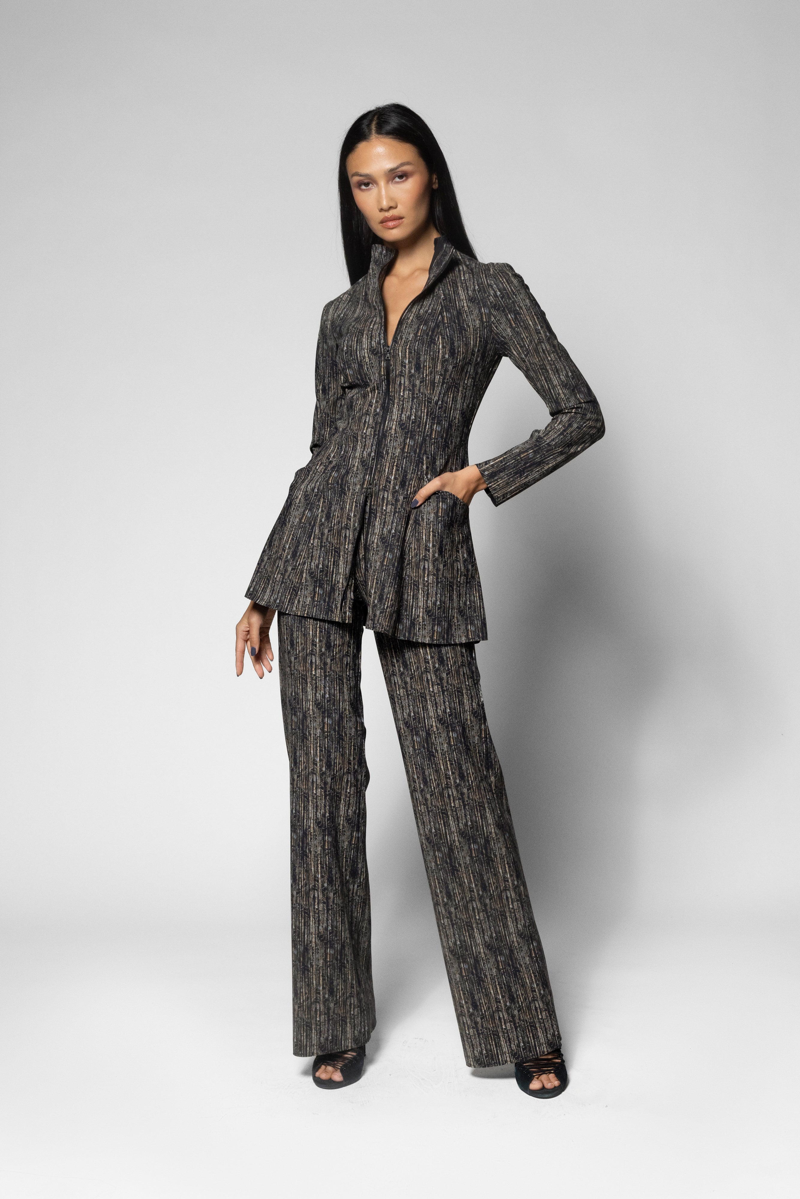 A model wearing a dark gray and black textured two-piece suit with a two way zip up jacket and matching wide-leg trousers, standing against a light gray studio background.