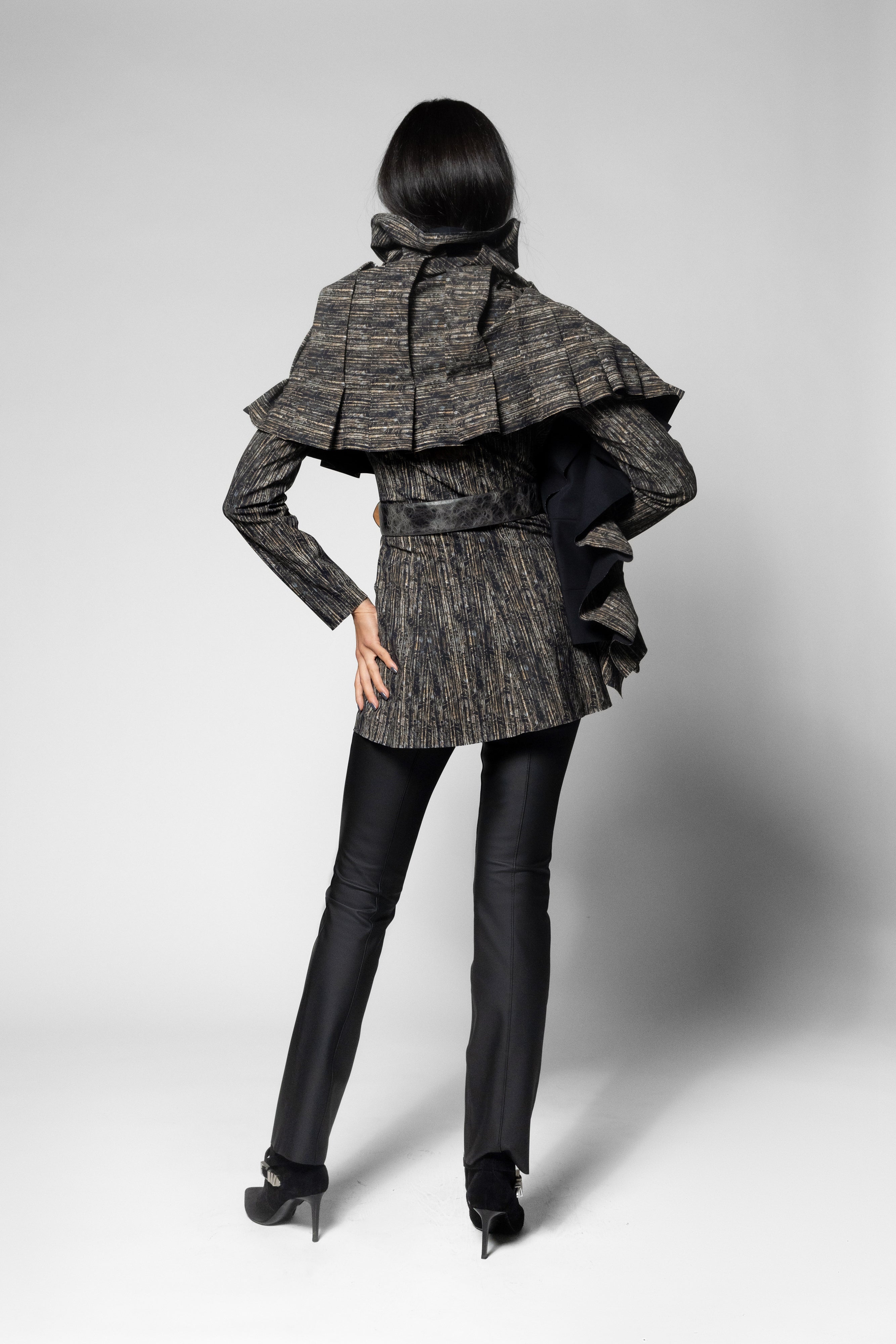 A model shown from the back wearing a textured, dark gray and black ruffled scarf over matching jacket with long sleeves, paired with black pants and black high heels, against a light gray studio background.