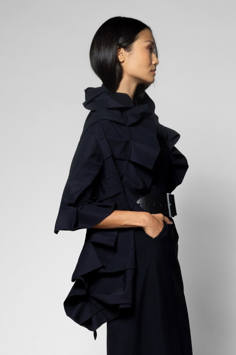 A model in profile wearing a black ruffled scarf with a high collar and a wide black belt, set against a plain light gray studio background.