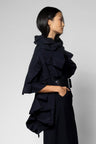 A model in profile wearing a black ruffled scarf with a high collar and a wide black belt, set against a plain light gray studio background.
