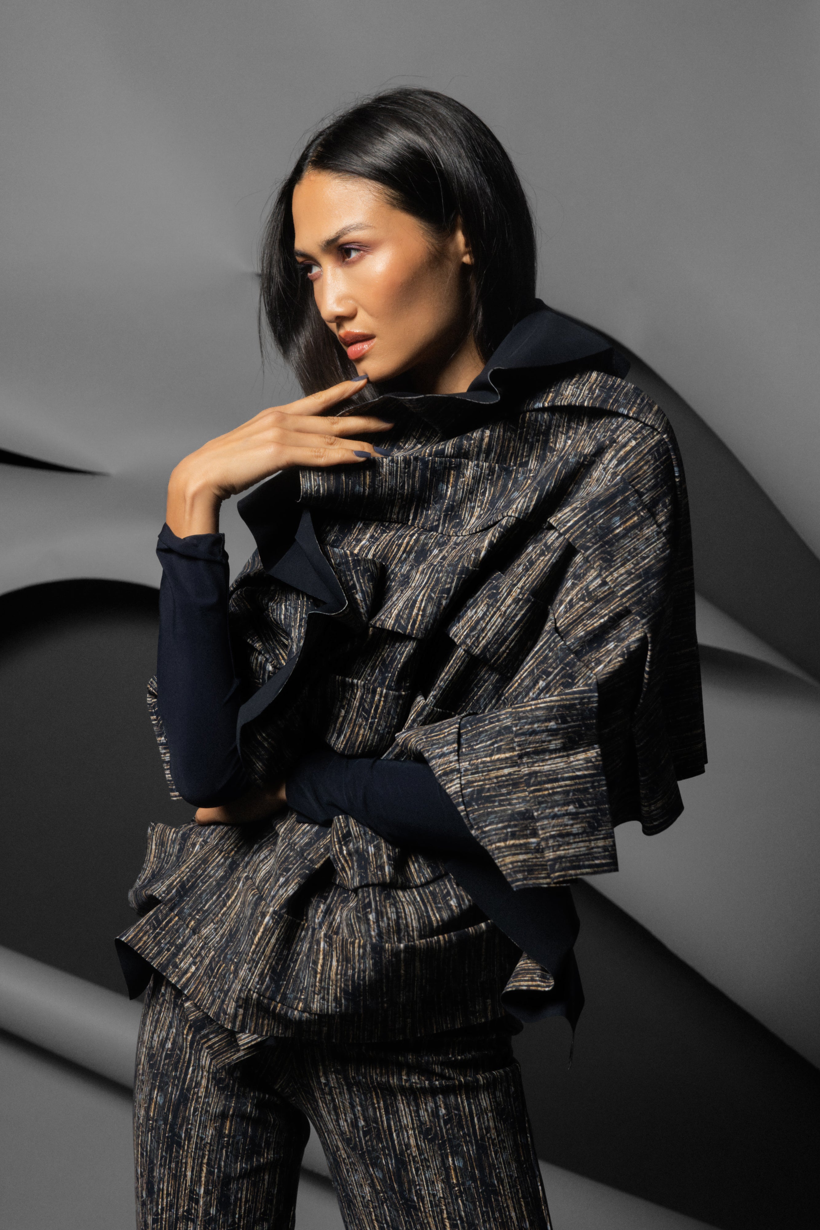 A model wearing a dark textured, shimmering ruffled scarf over a jacket with matching trousers and a black long-sleeve top, posed against a gray abstract studio background.