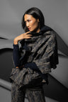 A model wearing a dark textured, shimmering ruffled scarf over a jacket with matching trousers and a black long-sleeve top, posed against a gray abstract studio background.
