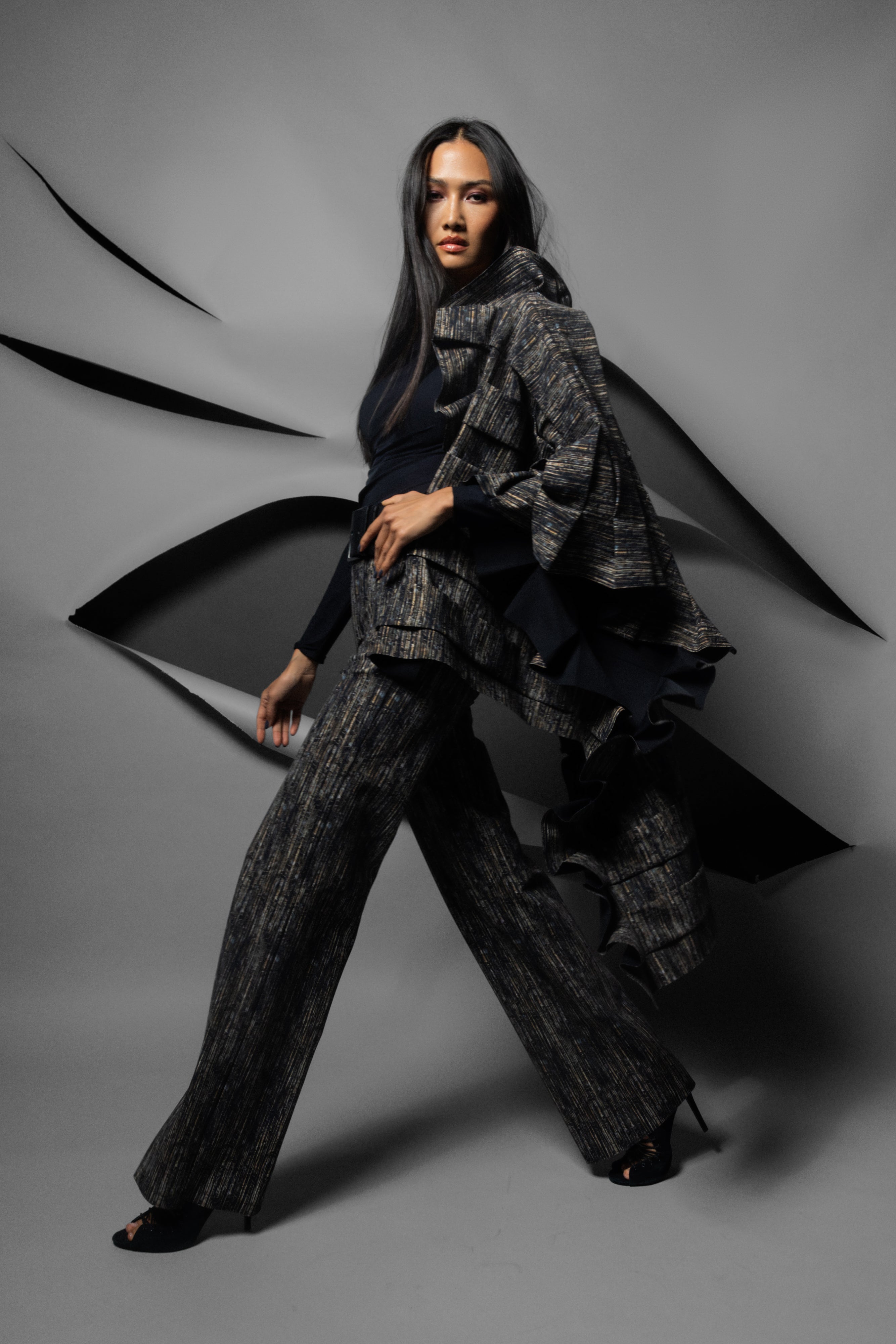 A model poses in a dark gray and black texture printed outfit consisting of wide-leg trousers and a matching ruffled scarf, set against a gray studio backdrop with abstract black paper-like shapes.