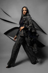 A model poses in a dark gray and black texture printed outfit consisting of wide-leg trousers and a matching ruffled scarf, set against a gray studio backdrop with abstract black paper-like shapes.