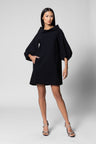 A female model wearing a black mini tunic with voluminous sleeves and a standing collar, paired with heels.