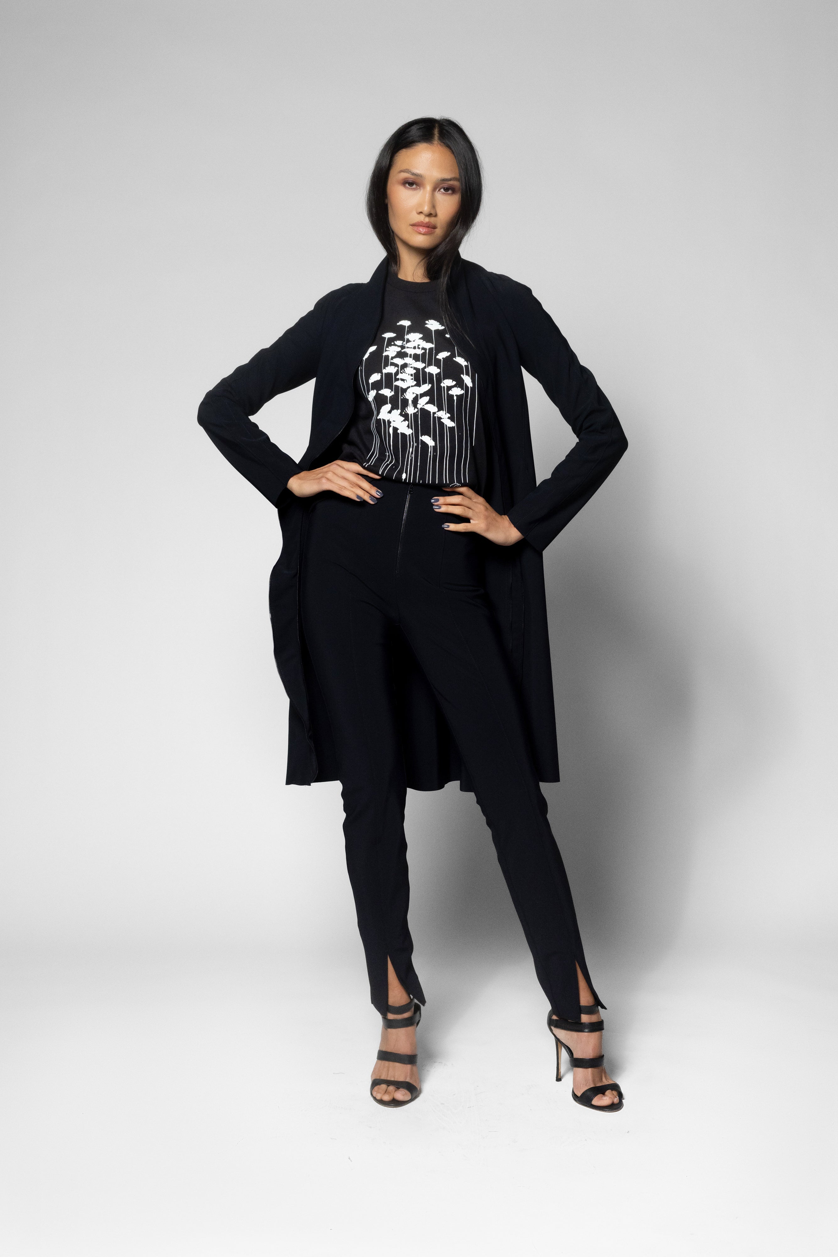 A model wearing a black long-sleeve jacket, black pants, and a black top with a white graphic print, standing against a light gray studio background.