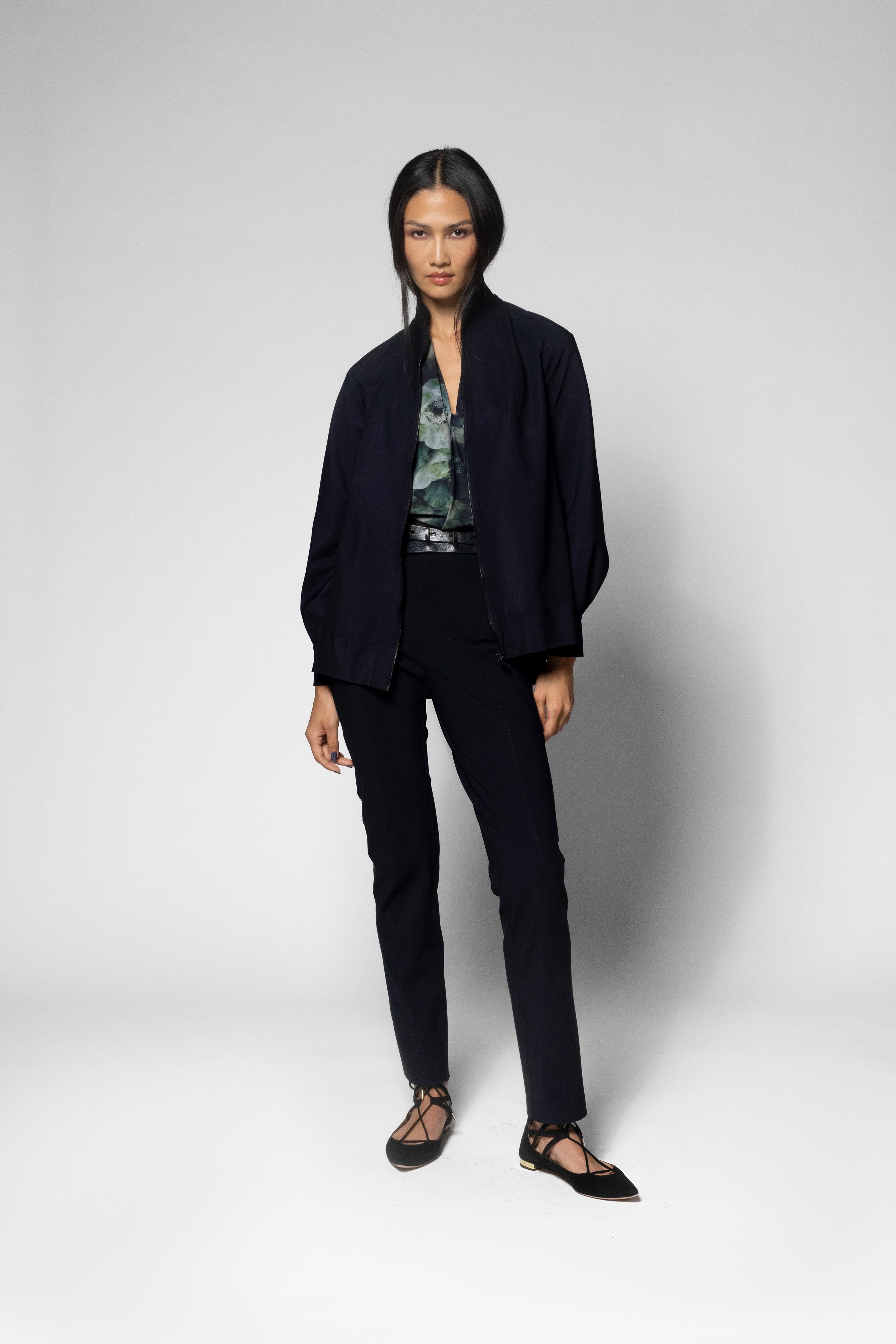 A model stands against a light gray studio background wearing a dark navy open-front jacket, matching dark trousers, a patterned blouse, and black strappy flat shoes.