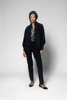 A model stands against a light gray studio background wearing a dark navy open-front jacket, matching dark trousers, a patterned blouse, and black strappy flat shoes.