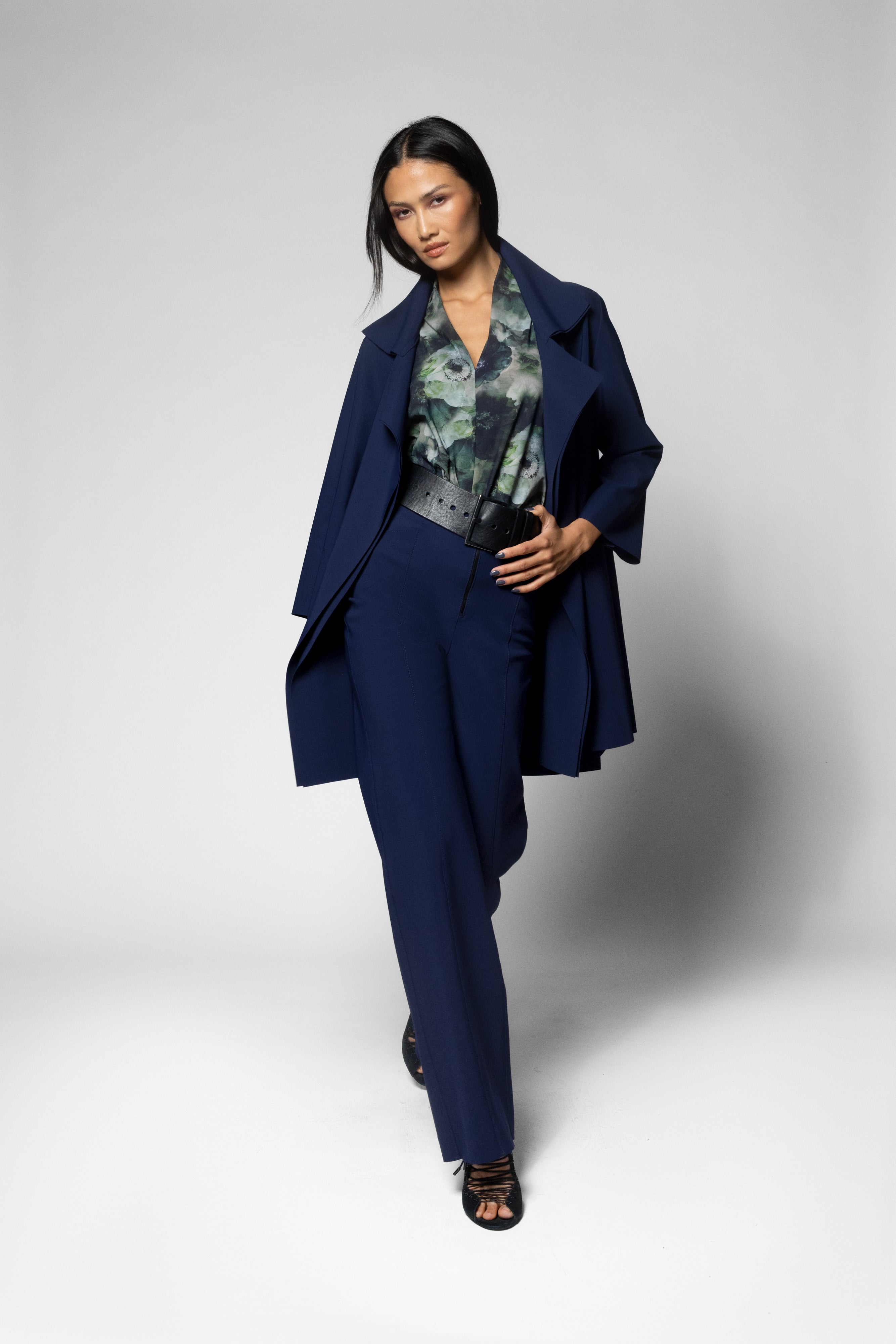 A model wearing a navy blue tailored suit consisting of a longline blazer and matching trousers, styled with a green and gray patterned blouse and dark heeled sandals, set against a light gray studio background.