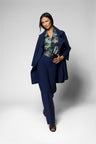 A model wearing a navy blue tailored suit consisting of a longline blazer and matching trousers, styled with a green and gray patterned blouse and dark heeled sandals, set against a light gray studio background.