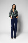 A woman stands against a light gray studio background wearing a dark green and black patterned blouse with three-quarter sleeves, a black belt, and navy blue straight-leg trousers.