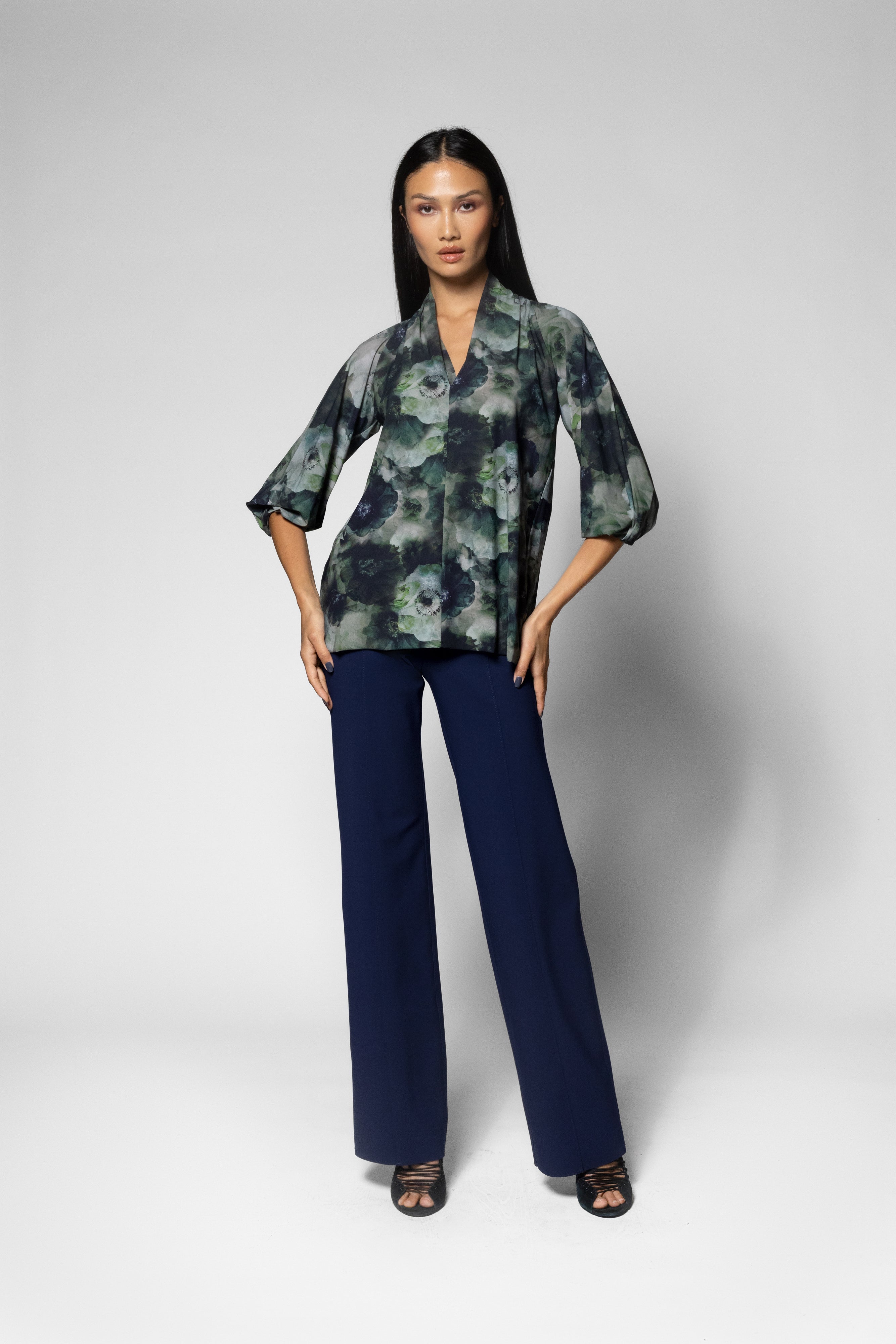 A woman modeling a 3/4 sleeve shirt with a green and black abstract floral pattern, paired with navy blue wide-leg trousers, against a light gray studio background.