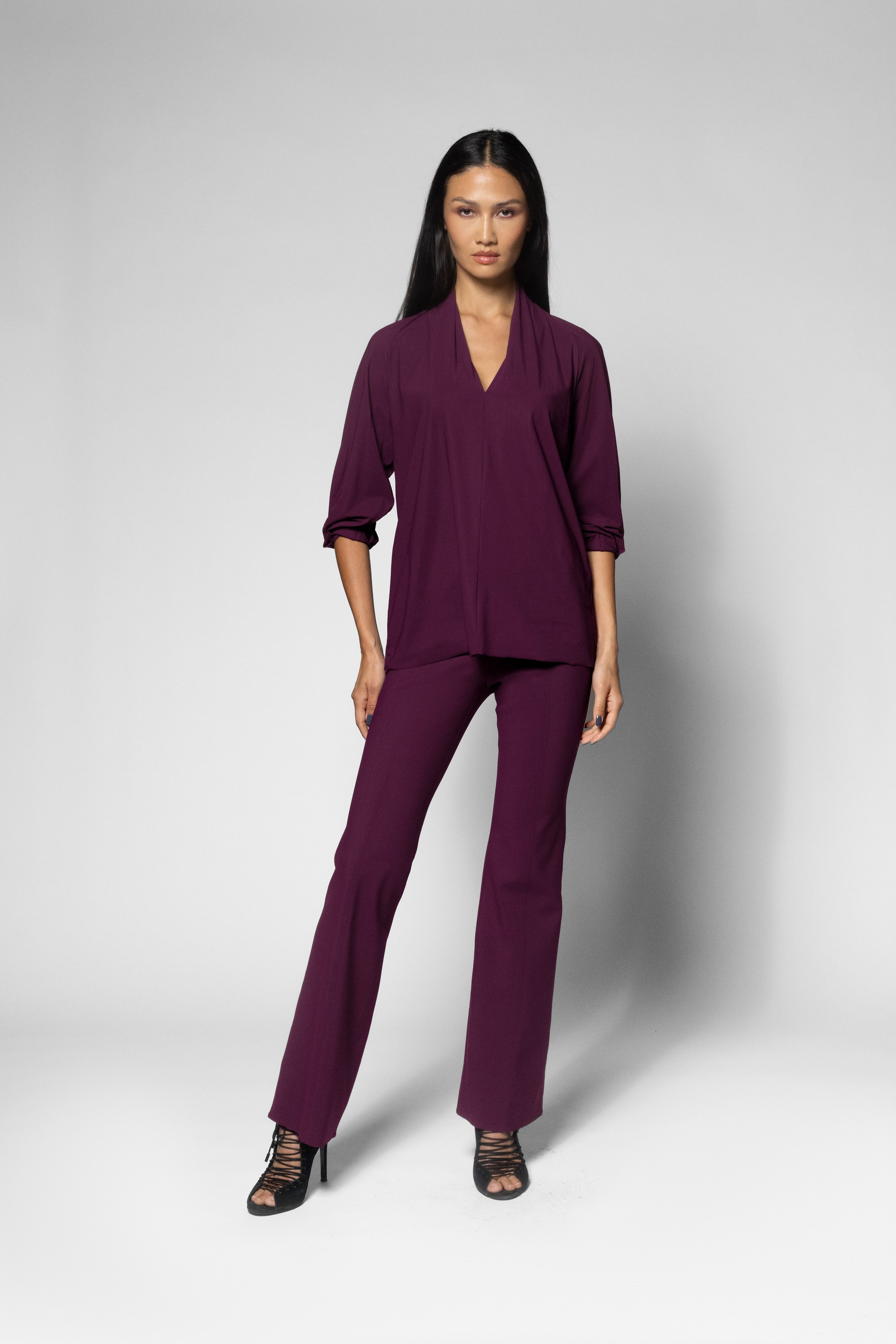 A model wearing a matching deep plum purple two-piece set consisting of a V-neck long-sleeve top and coordinating wide-leg pants, paired with black strappy heels, against a light gray studio background.