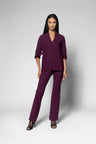 A model wearing a matching deep plum purple two-piece set consisting of a V-neck long-sleeve top and coordinating wide-leg pants, paired with black strappy heels, against a light gray studio background.