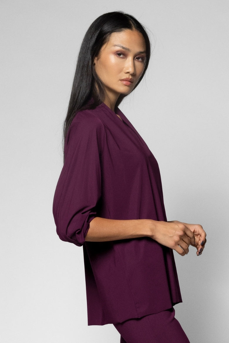 A woman modeling a deep plum purple three-quarter sleeve top and matching pants against a light gray studio background.