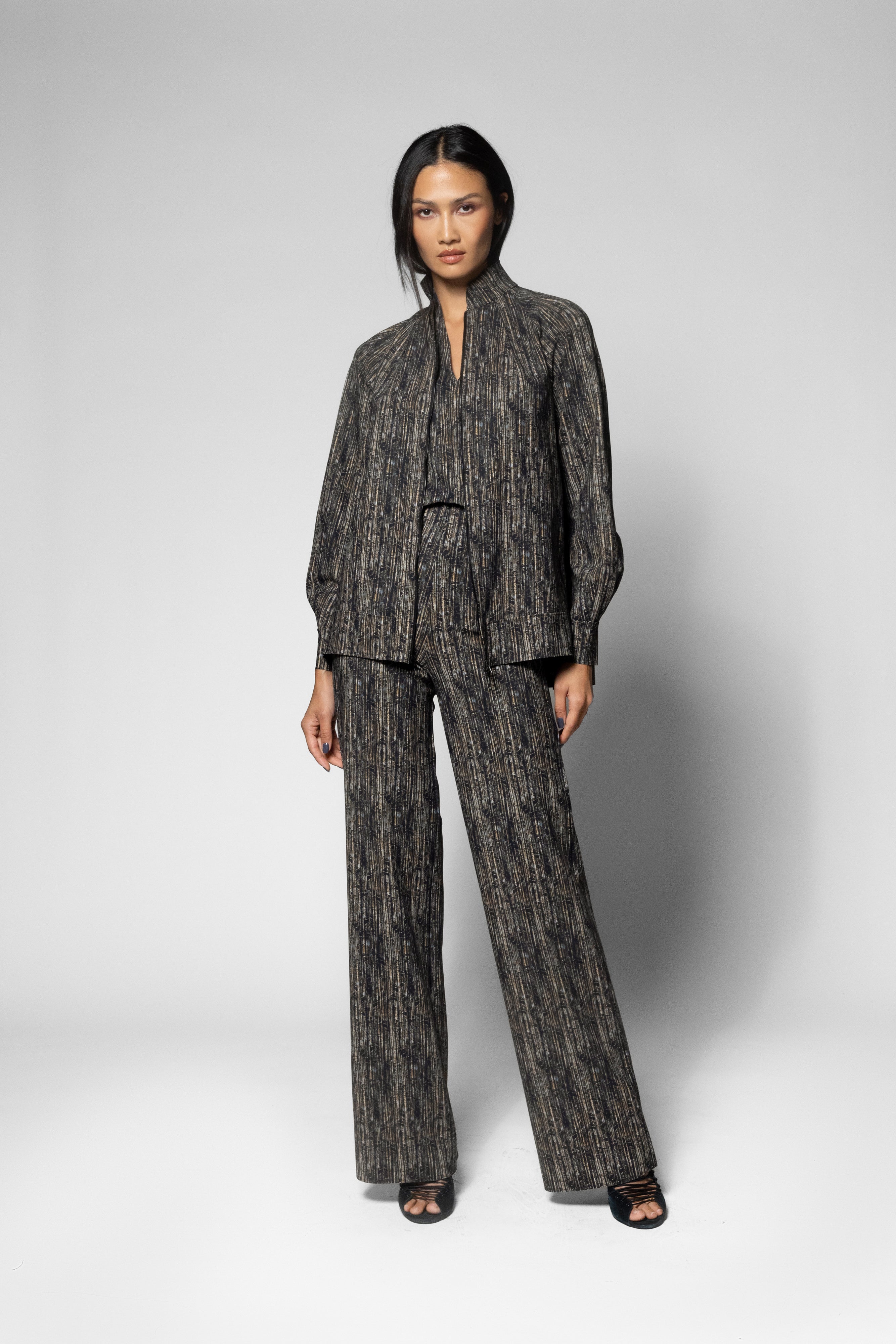 A model stands against a light gray studio background wearing a matching dark gray and black patterned two-piece suit with a long-sleeve jacket and wide-leg trousers.