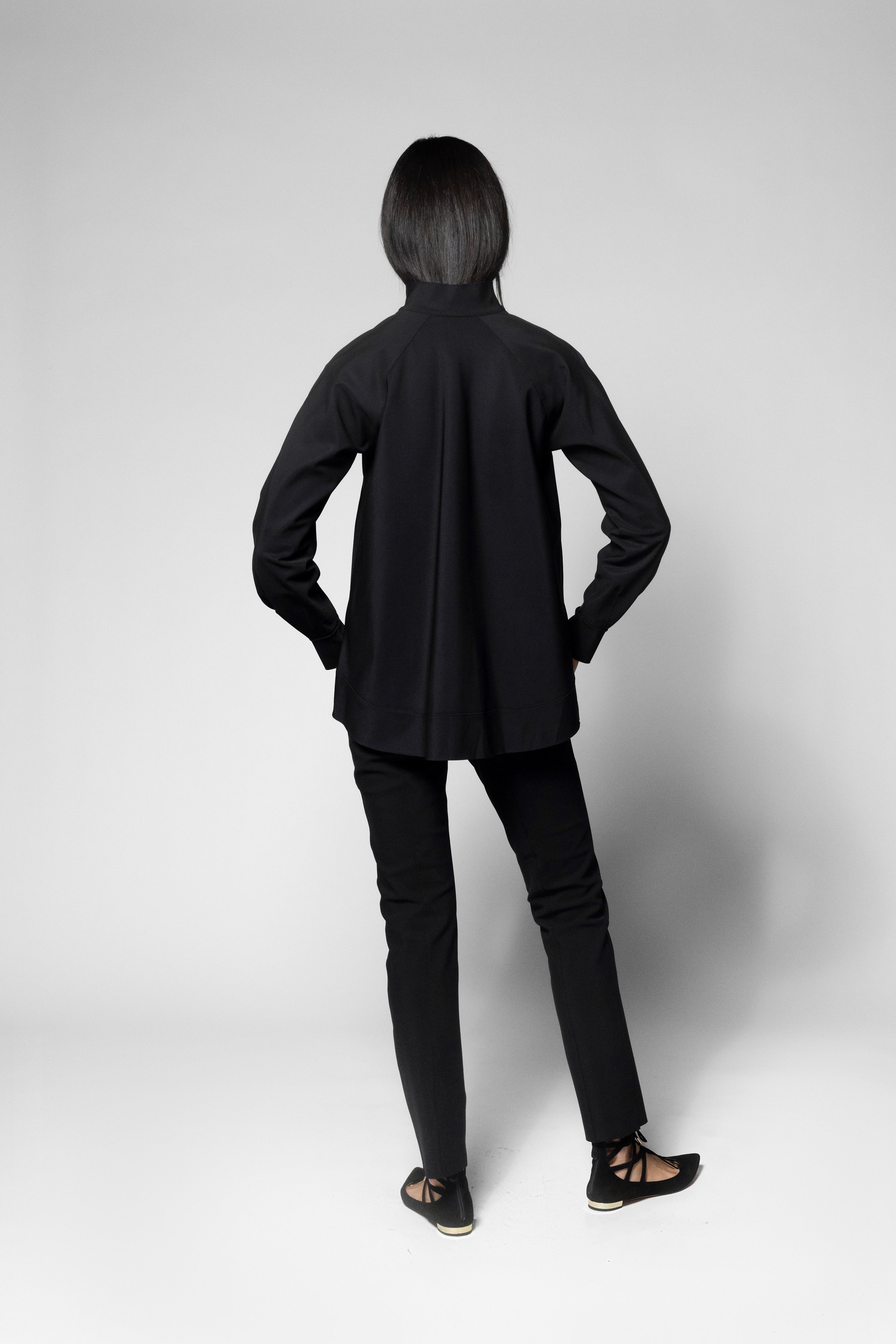 A model viewed from the back wearing a black long-sleeve mock-neck jacket, black slim-fit pants, and black strappy flat sandals against a light gray studio background.