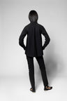 A model viewed from the back wearing a black long-sleeve mock-neck jacket, black slim-fit pants, and black strappy flat sandals against a light gray studio background.