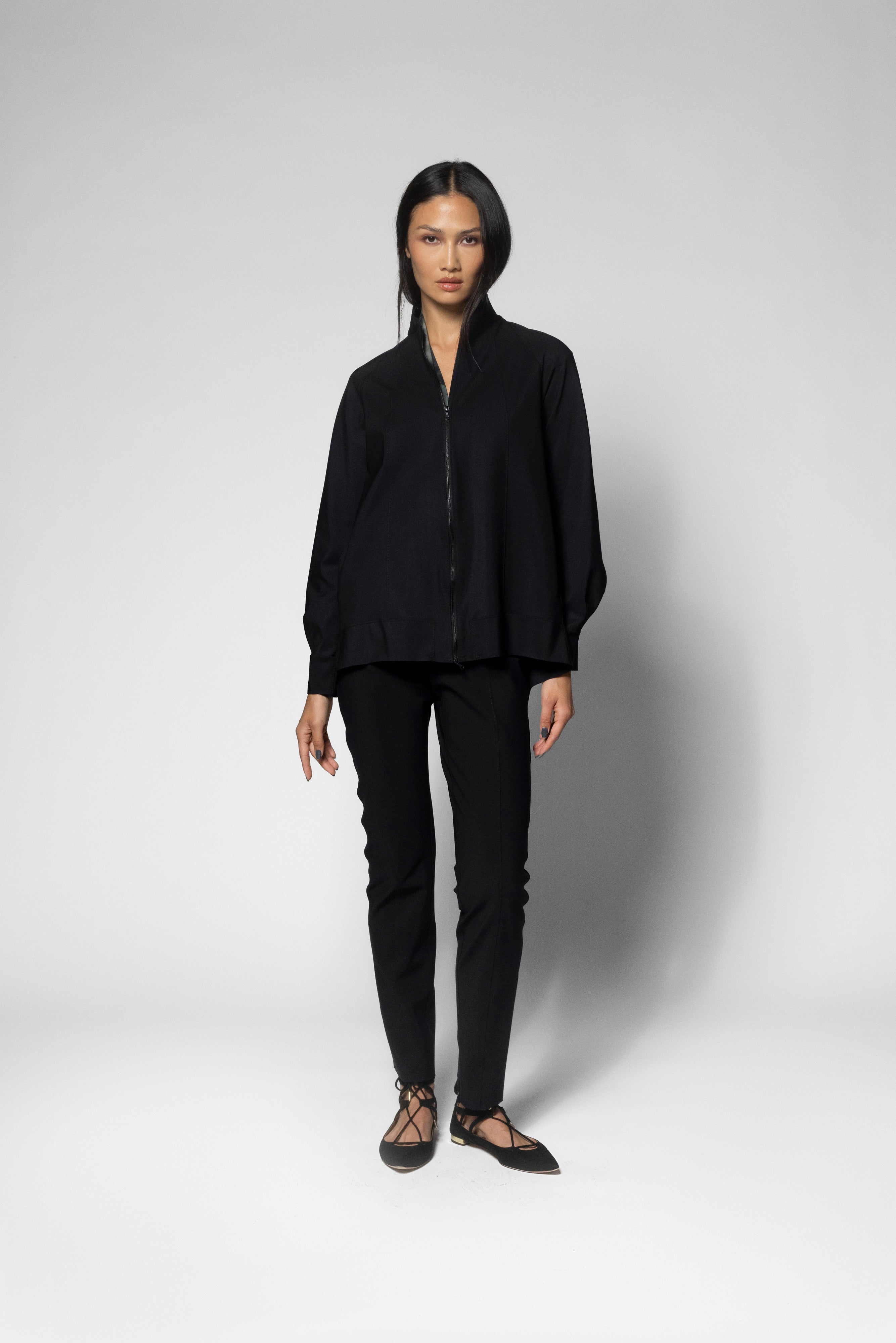 A model stands against a light gray studio background wearing a black zip-front jacket, black slim-fit pants, and black lace-up flats.