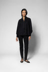 A model stands against a light gray studio background wearing a black zip-front jacket, black slim-fit pants, and black lace-up flats.