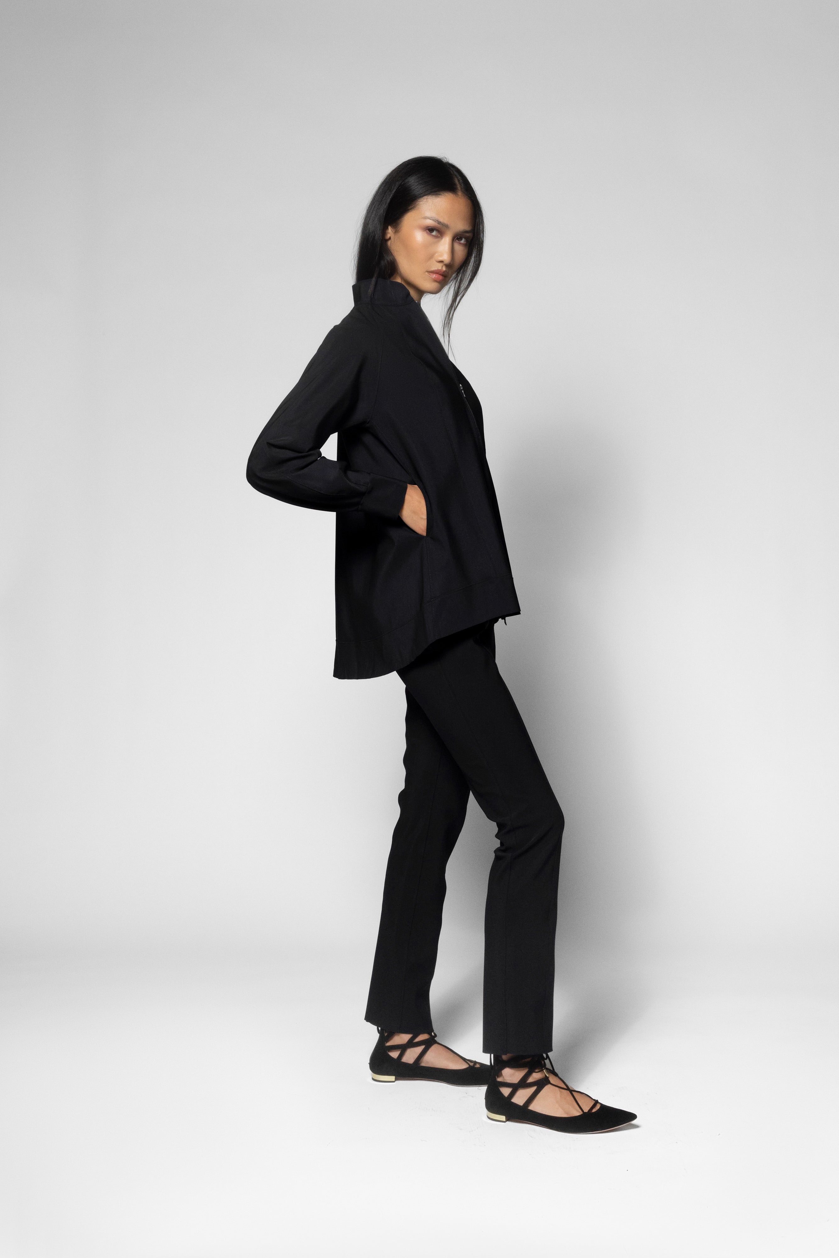 A woman modeling a black tailored jacket and matching black trousers, paired with black strappy flat shoes, against a light gray studio background.
