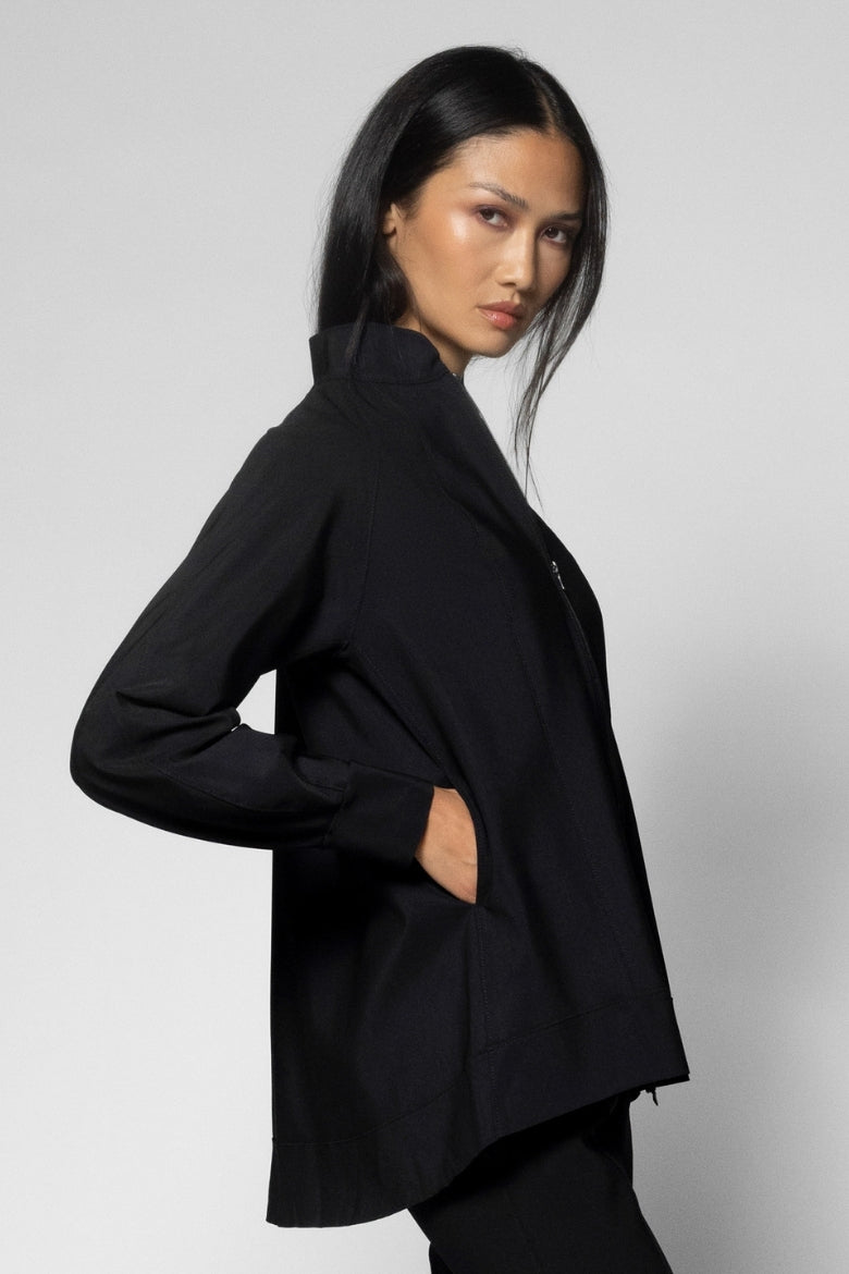 A woman modeling a black long-sleeve jacket with a high stand collar, shown from a three-quarters back angle against a light gray studio background.