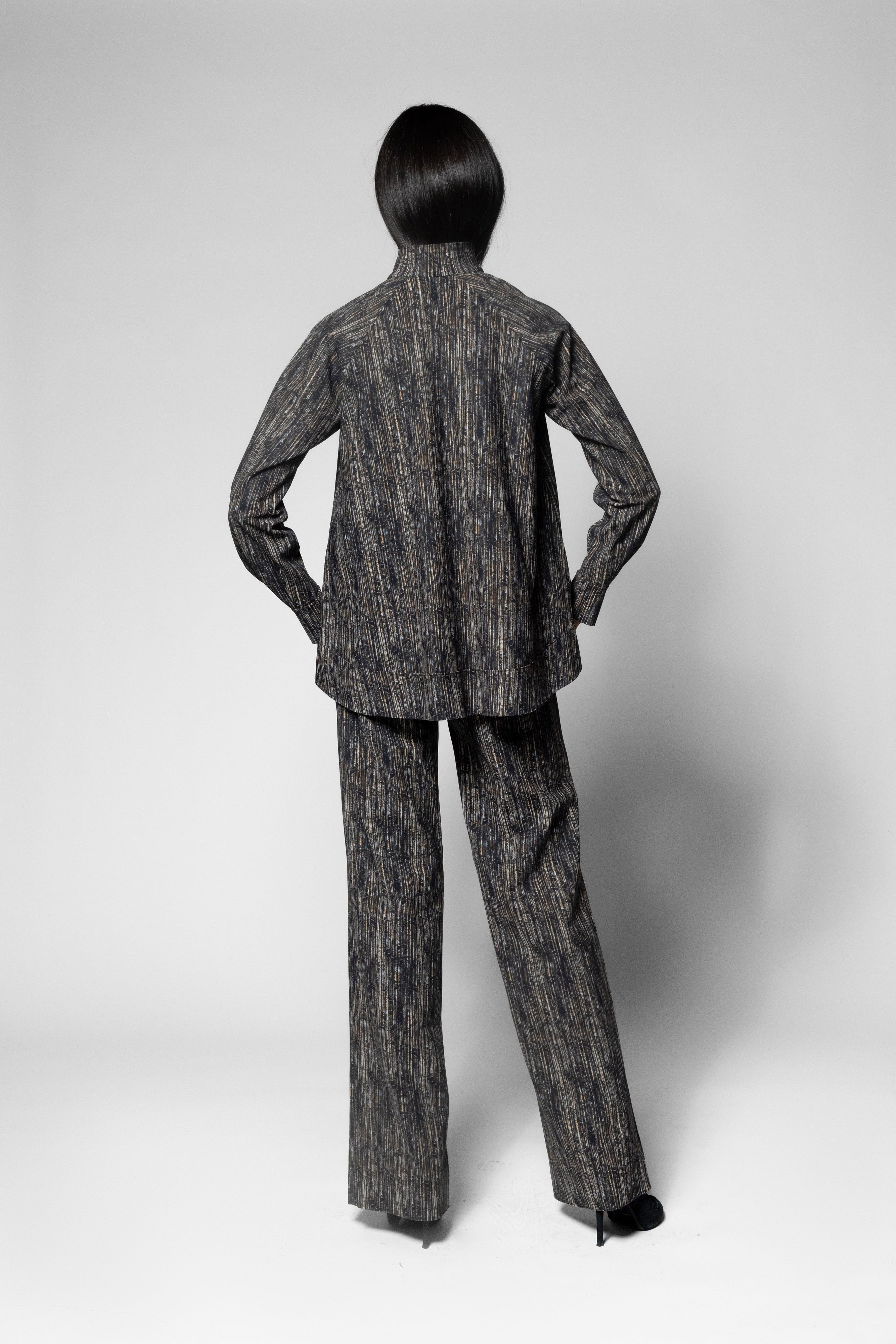 A model shown from the back wearing a matching dark gray and black textured patterned blazer and trousers set, photographed against a neutral light gray studio background.