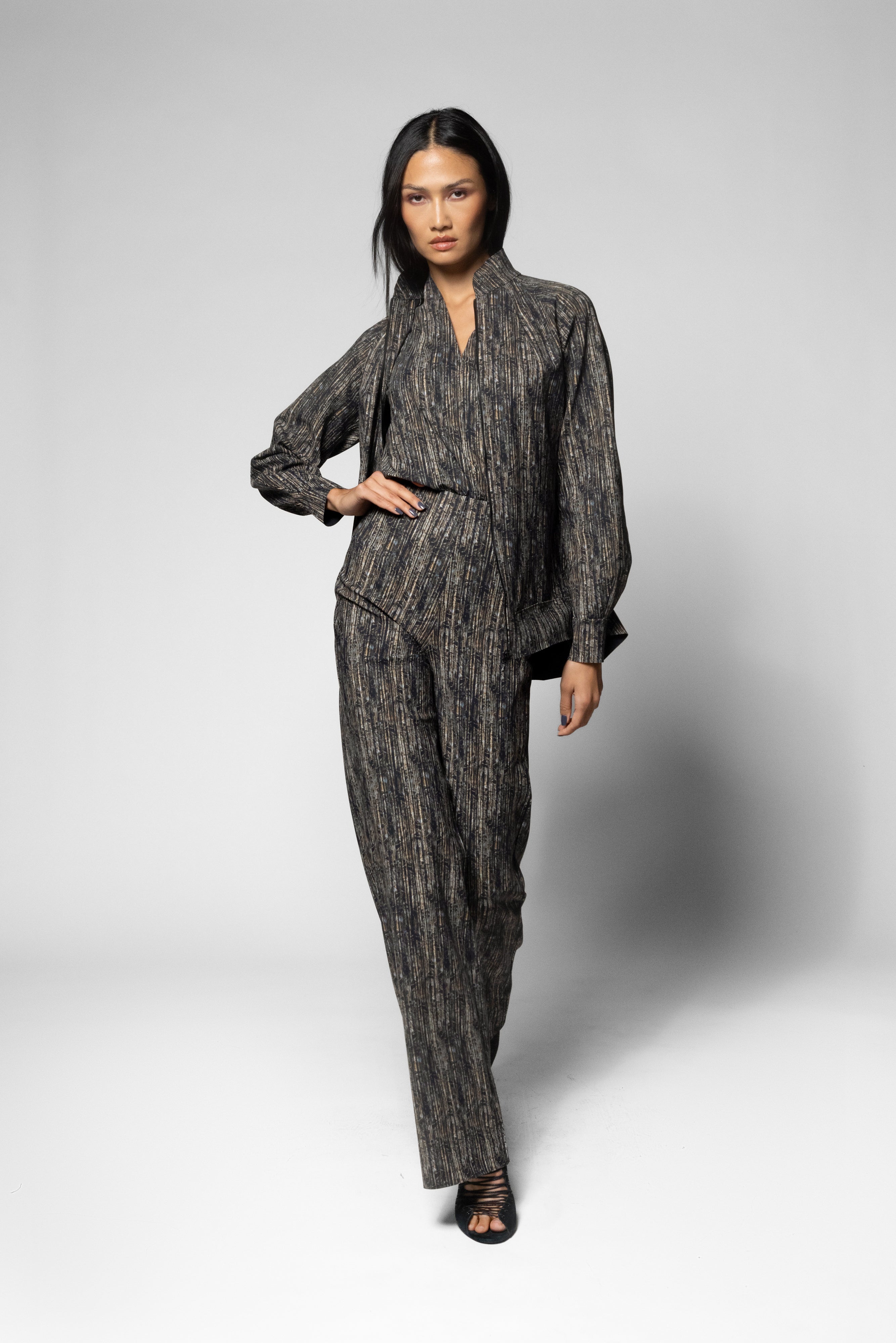 A model walks against a light gray studio background wearing a dark gray and black textured, vertically-striped two-piece set consisting of a long-sleeve collared shirt and matching trousers.