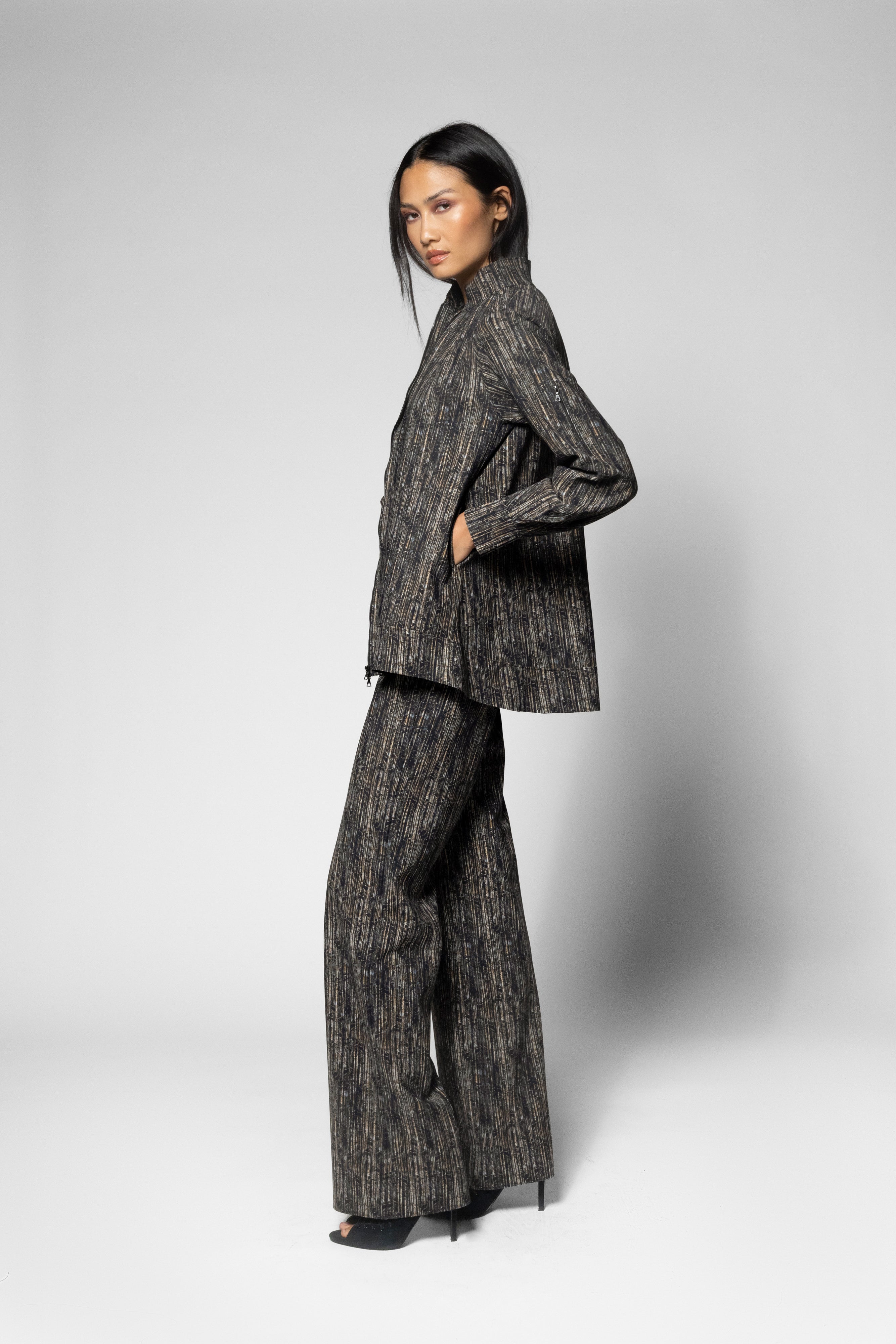 A model wearing a matching textured gray and black jacket and wide-leg trousers suit, standing in profile against a light gray studio background.