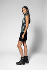 A woman modeling a sleeveless top with a green and black abstract pattern, paired with a black skirt and black combat boots, standing against a light gray studio background.