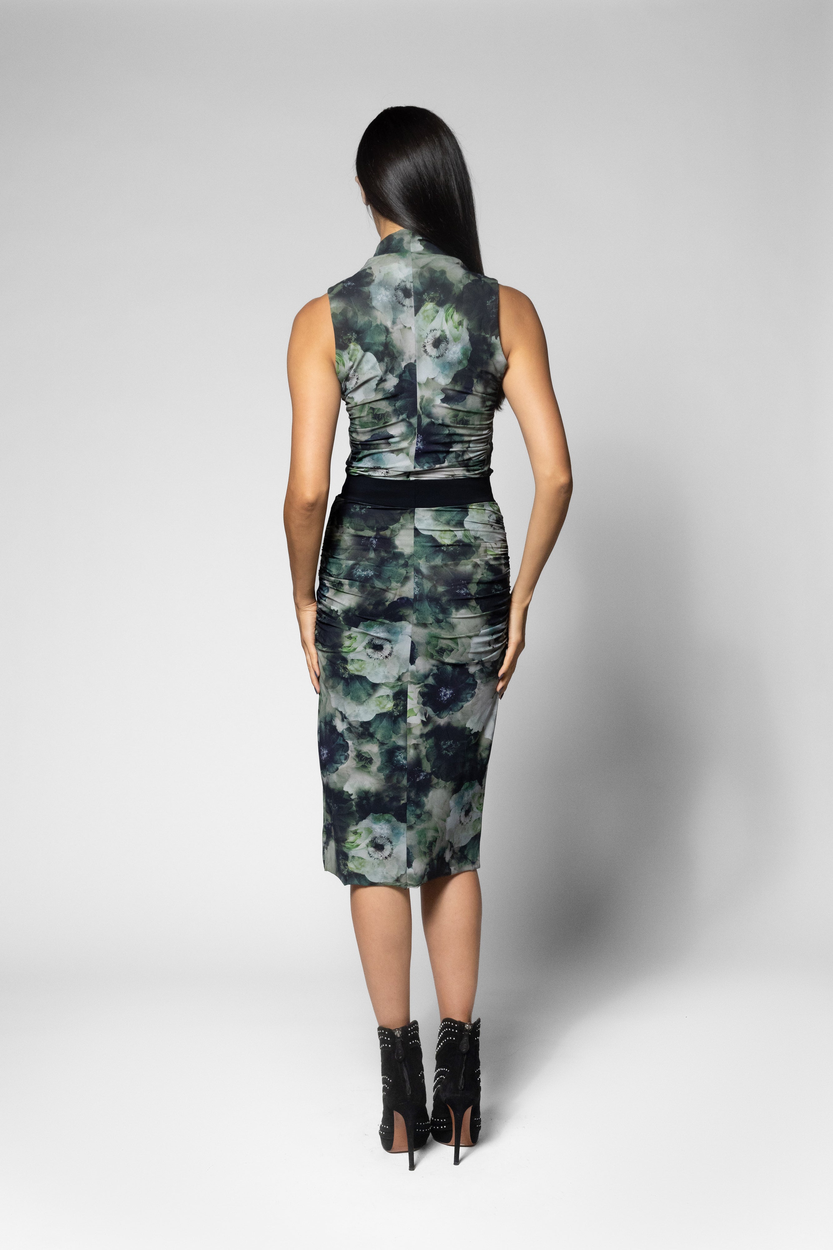 A woman shown from the back wearing a sleeveless top and knee-length skirt with a dark floral print in green, gray, and black tones, paired with black ankle boots, against a light gray studio background.