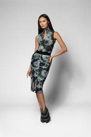 A model wearing a sleeveless high-neck shirt and midi skirt with a green and black abstract floral pattern, paired with black ankle boots, against a light gray studio background.