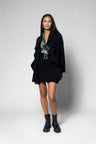 A model stands against a light gray studio background wearing a black asymmetrical draped jacket, a black mini skirt with a silver belt, a patterned top, and black combat boots.