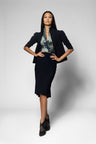 A woman modeling a black two-piece skirt suit consisting of a cropped blazer and a matching knee-length skirt, worn over a green and blue patterned blouse with black pointed-toe heels, against a light gray studio background.