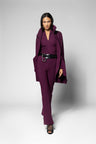 A model wearing a deep plum purple three-piece suit consisting of a collared long-sleeve top, matching trousers, and a wide black belt with a silver buckle, standing against a light gray studio background.